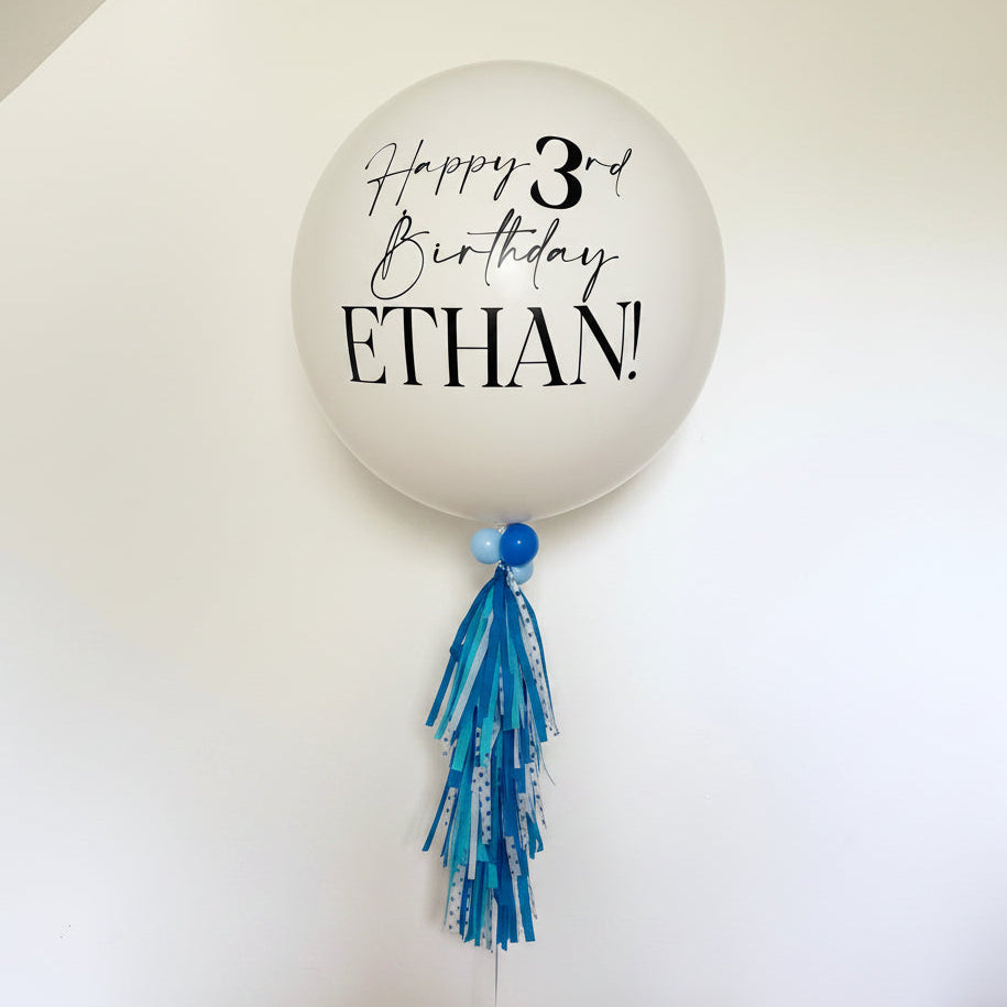 A jumbo white latex balloon with a personalized message 'Happy 3rd Birthday Ethan' printed on it, adorned with a blue tassel.